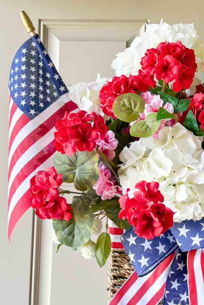 Close up of a patriotic summer door basket with red geraniums, white hydrangeas, and pink flowers in a woven basket tied with a stars and stripes ribbon bow and a small American flag