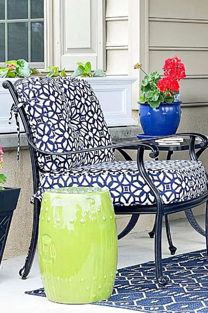 Summer porch with black iron patio chairs with navy and white geometric cushions, a matching outdoor rug, a blue pot of red geraniums, and a white ceramic planter on a side table overlooking a green landscape