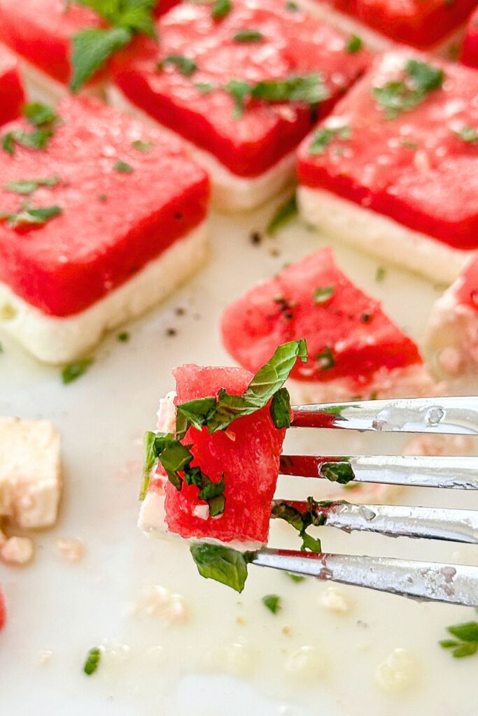 Close up of a fork holding a bite of watermelon and feta salad with fresh mint over a platter of stacked watermelon and feta squares for a summer Memorial Day recipe