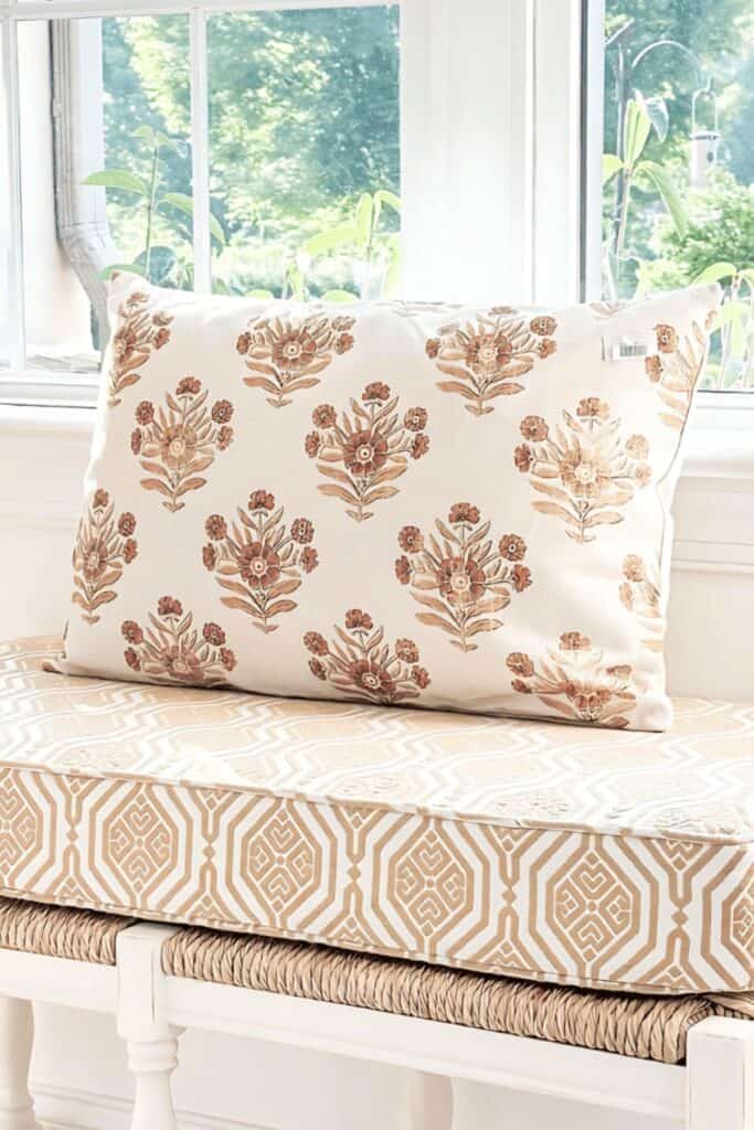 White bench with a woven seat, a gold and white geometric print cushion, and a large cream and rust block print floral pillow in front of bright windows with summer greenery outside