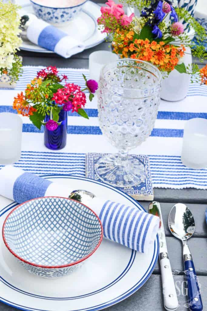 blue and white al fresco dining table setting with hobnail glasses, colorful summer flowers, and striped napkins