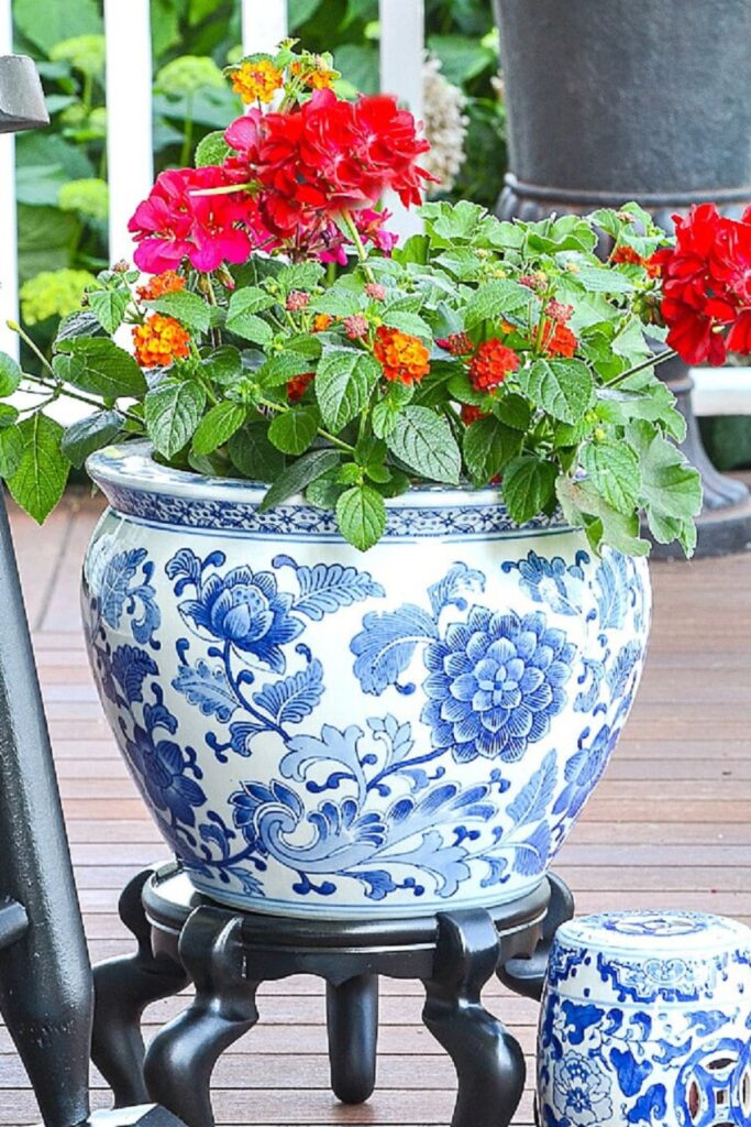Large blue and white chinoiserie fishbowl planter filled with red geraniums and orange lantana on a black wooden stand on a deck with a matching garden stool nearby