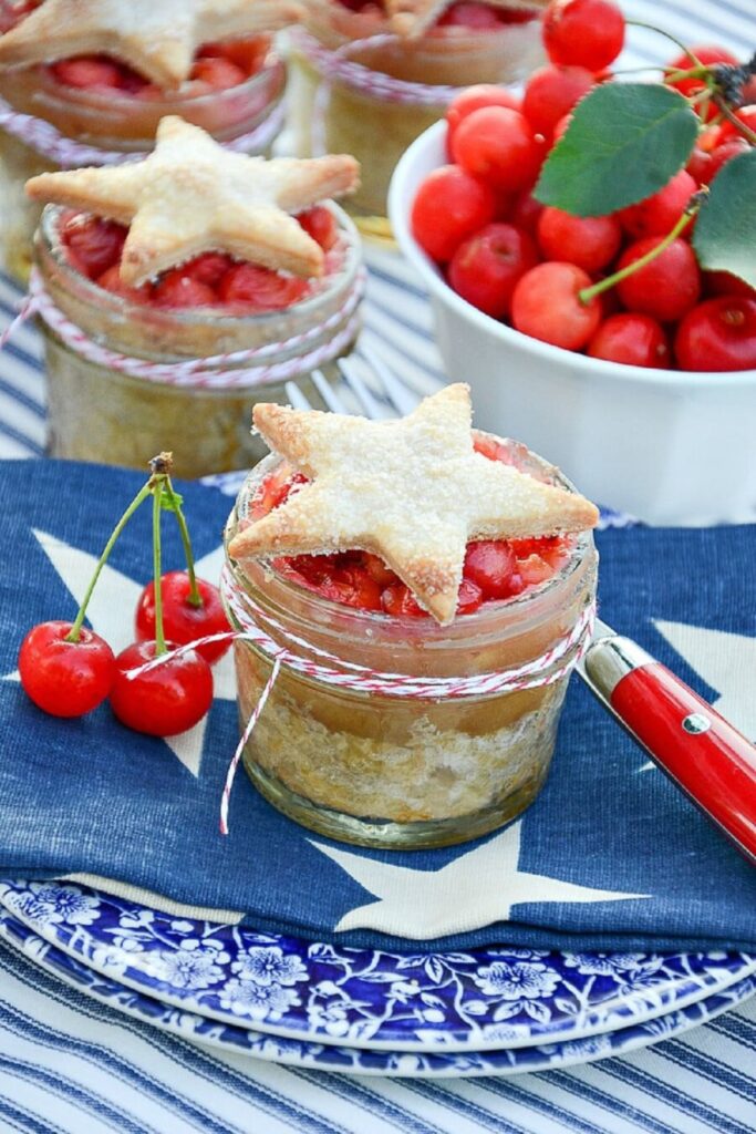 Individual cherry pies baked in mason jars with star shaped pie crust tops tied with red and white baker's twine on a blue and white star napkin and china plate with fresh cherries alongside