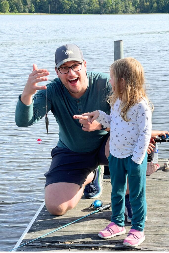 Laughing dad kneeling on a wooden dock holding up a small fish while his young daughter points at it on a lake during a Memorial Day weekend family fishing trip
