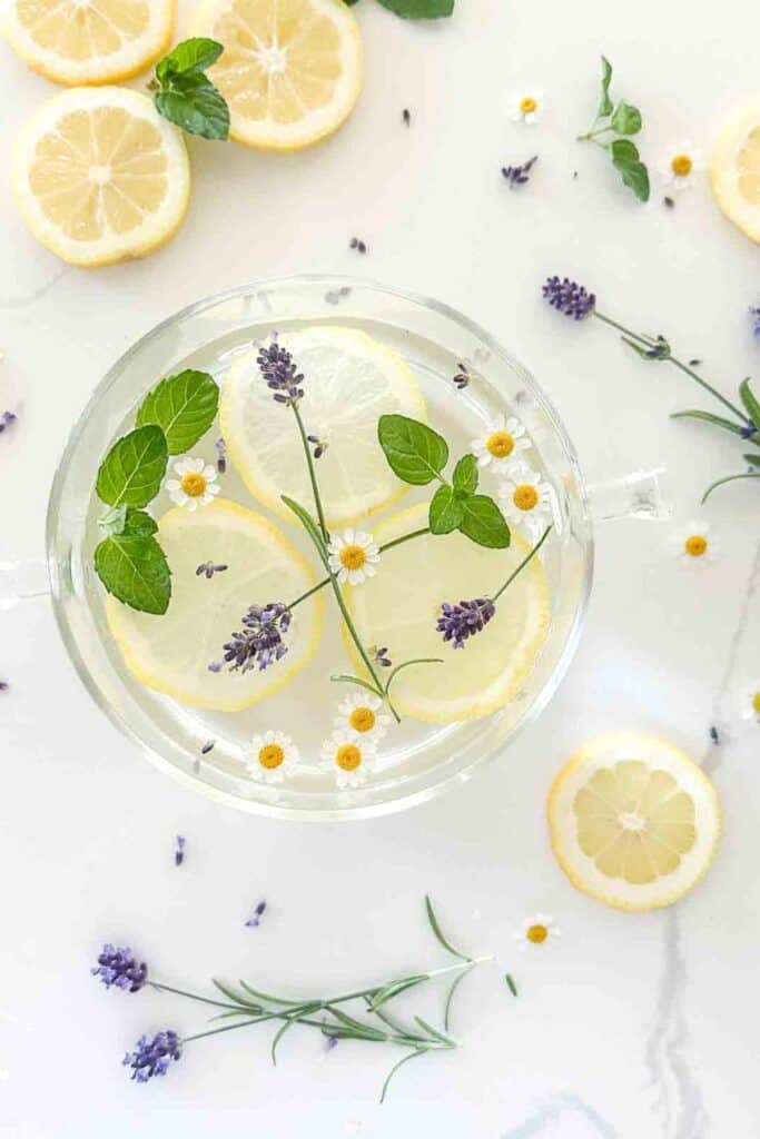 Overhead view of a glass pot of summer simmer pot with lemon slices, fresh mint leaves, lavender sprigs, and chamomile flowers on a white marble surface surrounded by scattered citrus and botanicals
