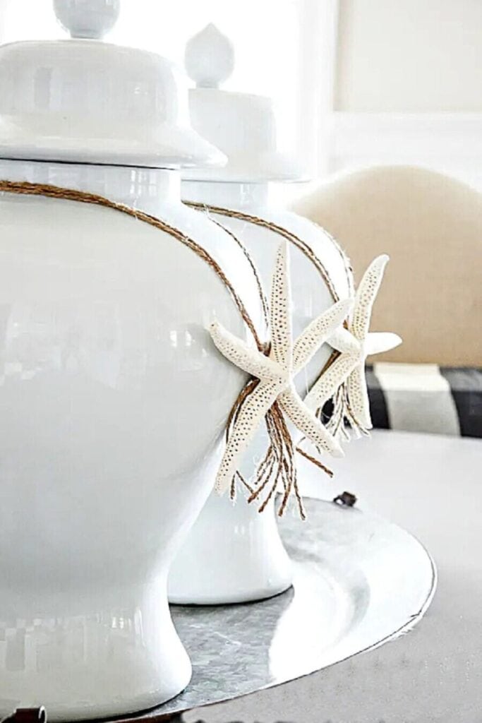 Two white ceramic ginger jars with white starfish tied around their necks with natural jute twine on a silver tray for a simple coastal summer accent