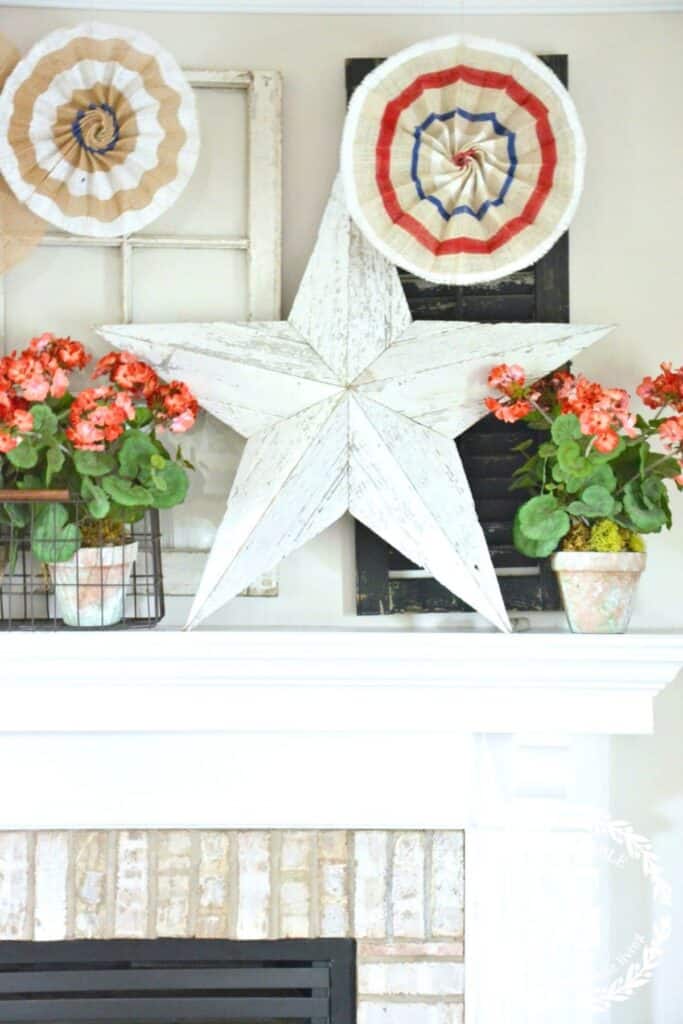 Patriotic mantel decorated with a large white chippy wood star, red geraniums in terra cotta pots, burlap rosette medallions, and a vintage black shutter