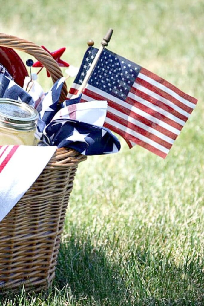 Wicker picnic basket sitting in green grass filled with a mason jar, red ticking stripe napkin, stars and stripes fabric, and a small American flag for a patriotic Memorial Day picnic
