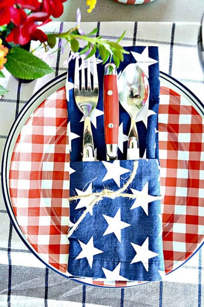 Patriotic place setting with a red gingham plate on a navy striped charger with red handled silverware tucked into a navy stars napkin tied with twine on a plaid tablecloth