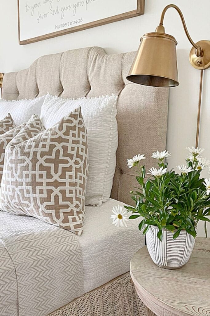 Neutral summer bedroom with a linen tufted headboard, a taupe geometric print pillow, white pom pom shams, a brass wall sconce, and a striped ceramic pot of white daisies on a round wood nightstand