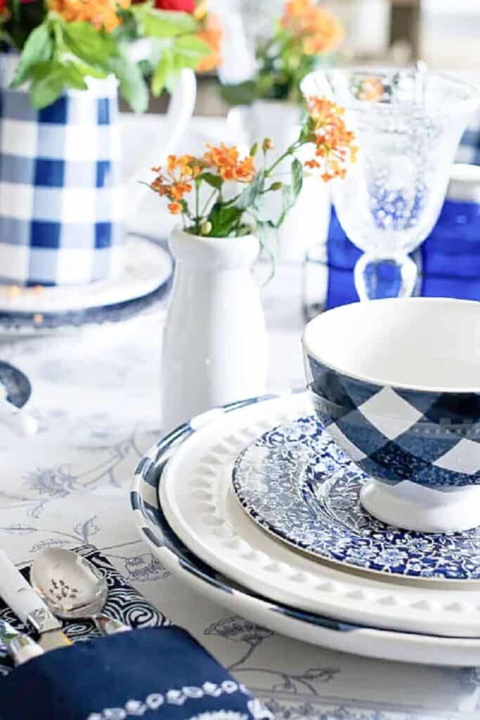 Close up of a layered blue and white summer table setting with mixed pattern dishes including a gingham bowl, floral salad plate, and beaded dinner plate, a small white pitcher of orange blooms, and a blue gingham tablecloth