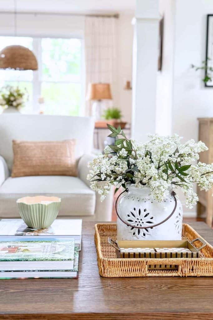 Summer coffee table vignette with a white pierced ceramic pot filled with white flowering branches and greenery in a woven rattan tray, a sage green ribbed bowl, and a stack of colorful books on a wood coffee table in a light filled living room