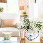 Pinterest graphic for 15 easy summer decorating ideas showing a light filled living room with a white pierced ceramic pot of white flowering branches in a woven tray on a coffee table with stacked books and a sage green bowl