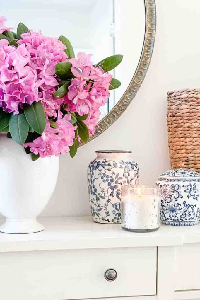 White foyer console table styled with a large white vase of pink rhododendron blooms, two blue and white chinoiserie ginger jars, a lit white candle, and a large woven basket tote beside a round mirror