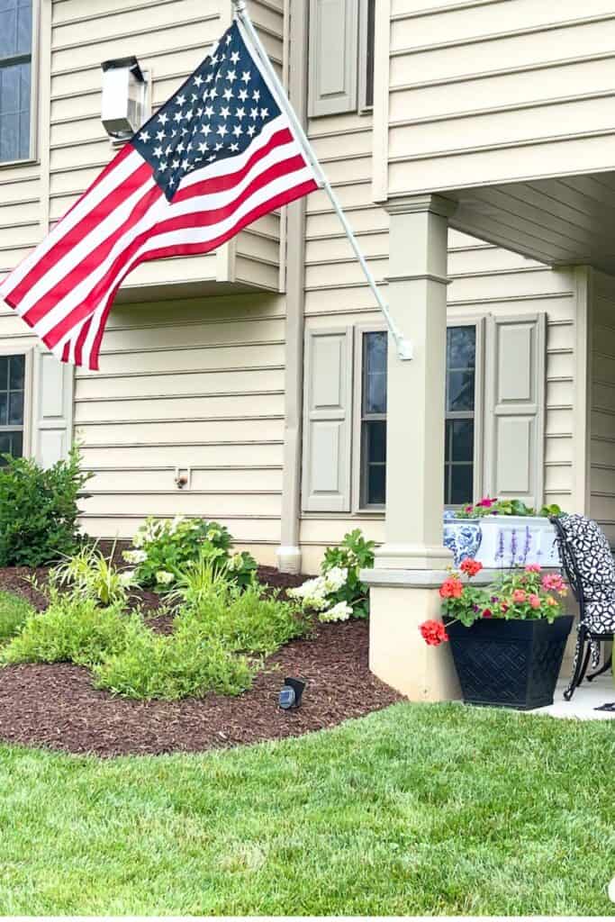 House exterior with an American flag flying from the front porch column, a black planter with red geraniums, a blue and white window box with summer flowers, and a well landscaped front garden bed