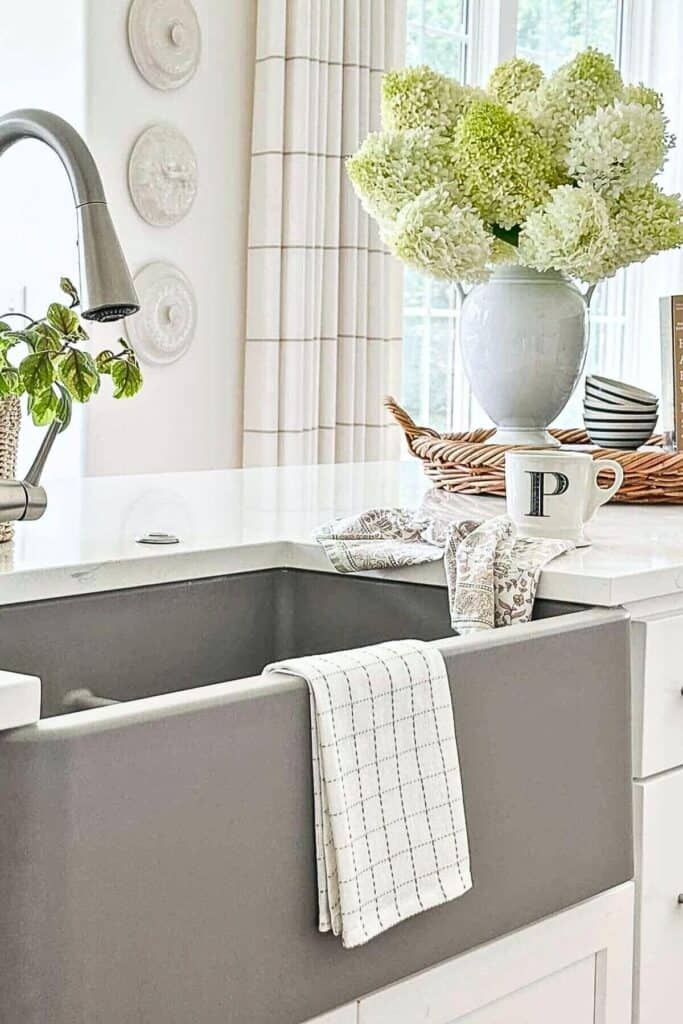 Gray farmhouse kitchen sink with a large white vase of green and white hydrangeas, a woven tray, striped bowls, and a monogram mug on a white quartz counter