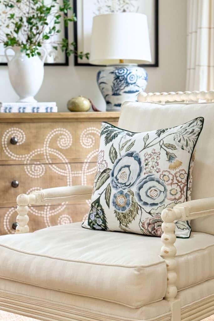 Cream spool chair with a blue and green botanical floral pillow, a carved wood dresser, blue and white ginger jar lamp, and a white pitcher of greenery in a summer living room vignette