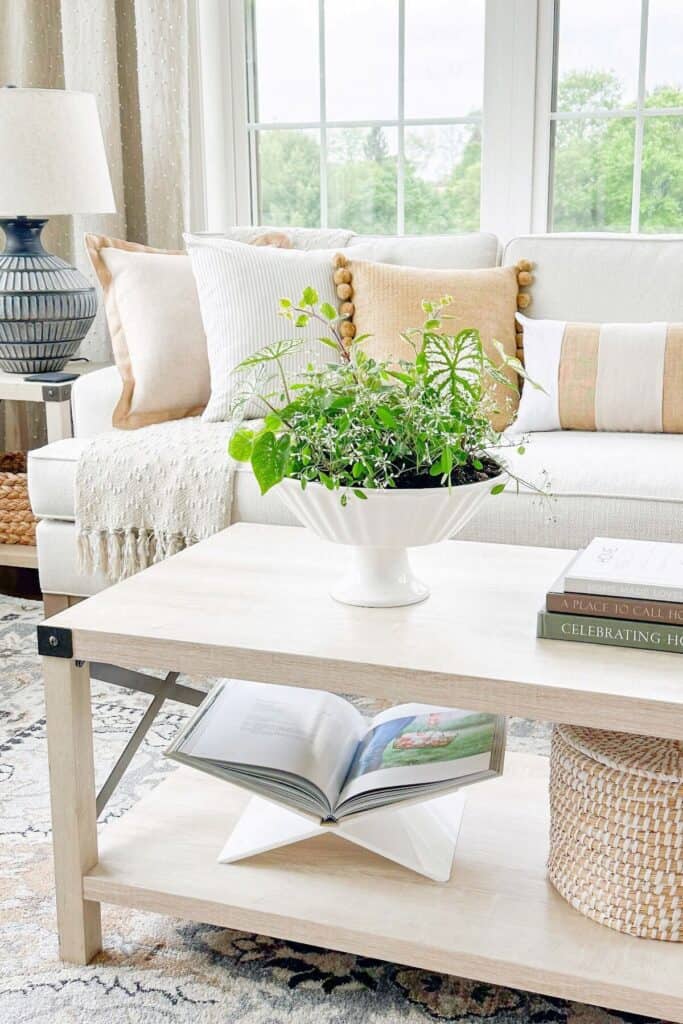 Light and airy living room with a white sofa styled with a jute pom pom pillow, a white striped pillow, and neutral linen pillows, a white coffee table with stacked books, a woven basket, and a potted green plant