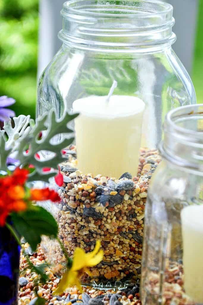 Close up of a clear mason jar filled with birdseed holding a white pillar candle as a summer outdoor lantern with colorful garden flowers and dusty miller in the background