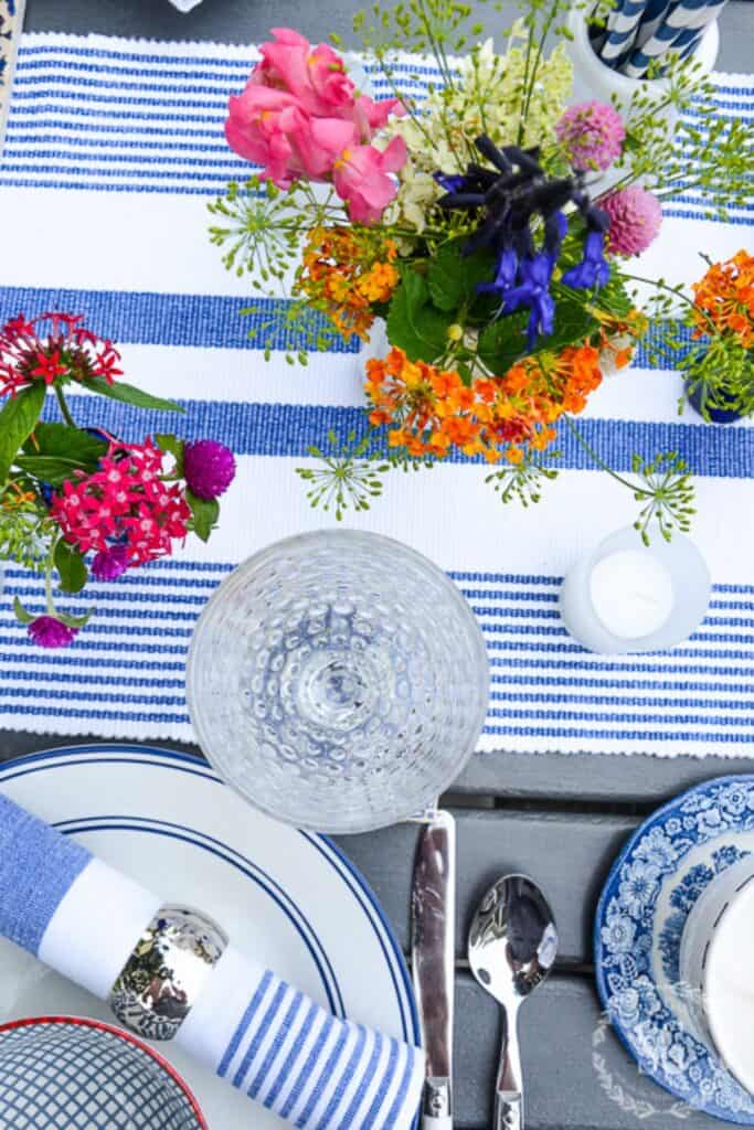 Overhead view of a colorful summer outdoor table setting with a blue and white striped runner, mixed blue and white dishes, crystal glassware, silver napkin rings, and a vibrant garden flower arrangement with pink roses, orange lantana, and purple blooms