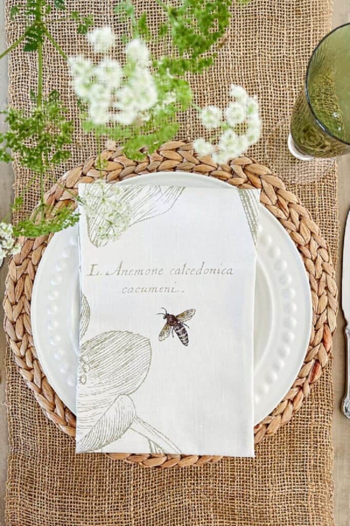 Overhead view of a summer place setting with a white botanical bee napkin on a white beaded plate, a woven rattan placemat, a burlap table runner, and small white wildflowers