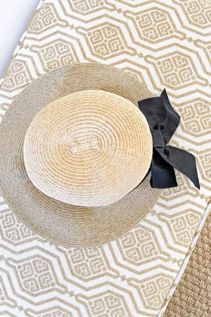 Overhead view of a straw sun hat with a black ribbon bow resting on a gold and white geometric patterned rug on a foyer bench