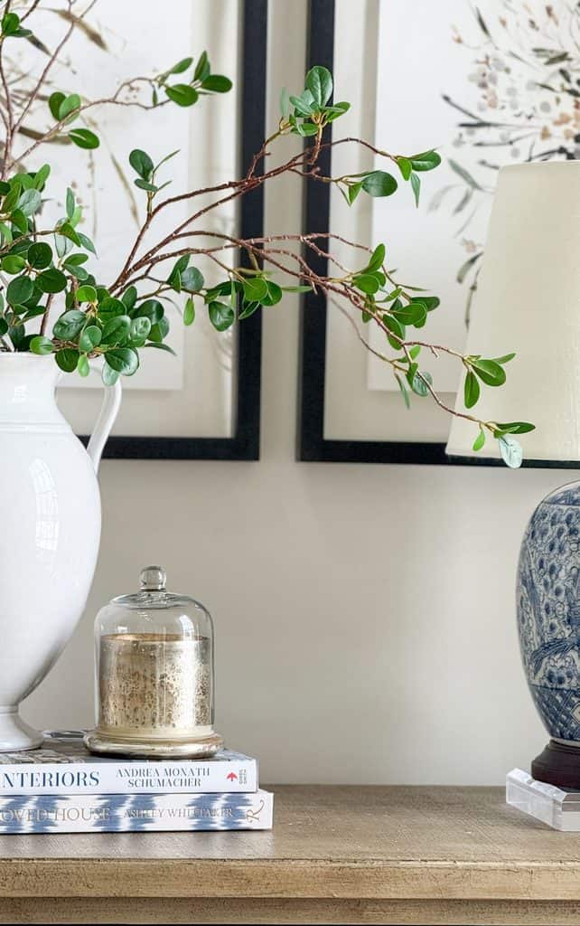 Console table vignette with a white pitcher of greenery, a mercury glass candle on stacked books, and a blue and white lamp to make a room prettier