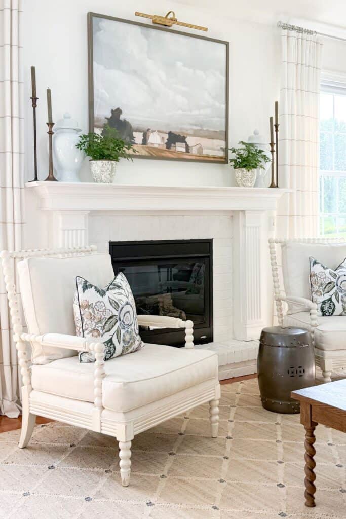white fireplace mantel with ginger jars and candlesticks, flanked by classic spindle chairs with floral pillows and a trendy metal garden stool