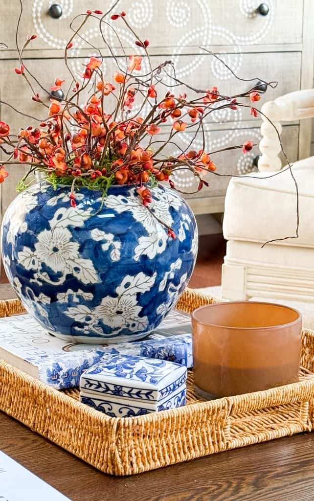 Blue and white vase with orange bittersweet stems on a woven tray with books, a blue and white box, and an amber candle to make a room prettier for fall