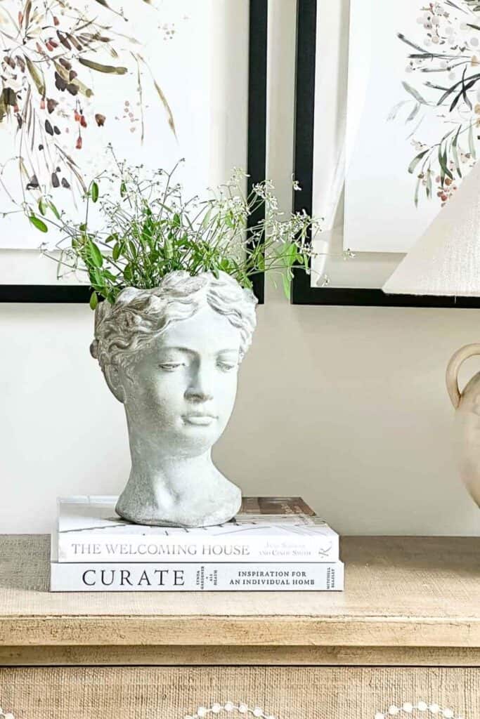 trendy stone goddess head planter with greenery, styled on a stack of books next to framed botanical art