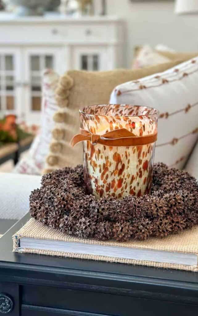 amber glass candle wrapped in a leather tie sitting inside a pinecone wreath on a burlap covered book