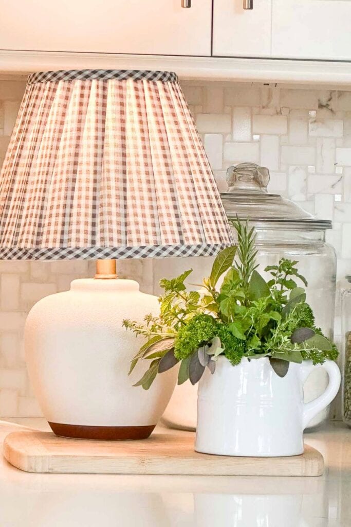 Soft white ceramic table lamp with a checkered fabric shade, styled with fresh greenery in vintage white pitcher on wooden tray.
