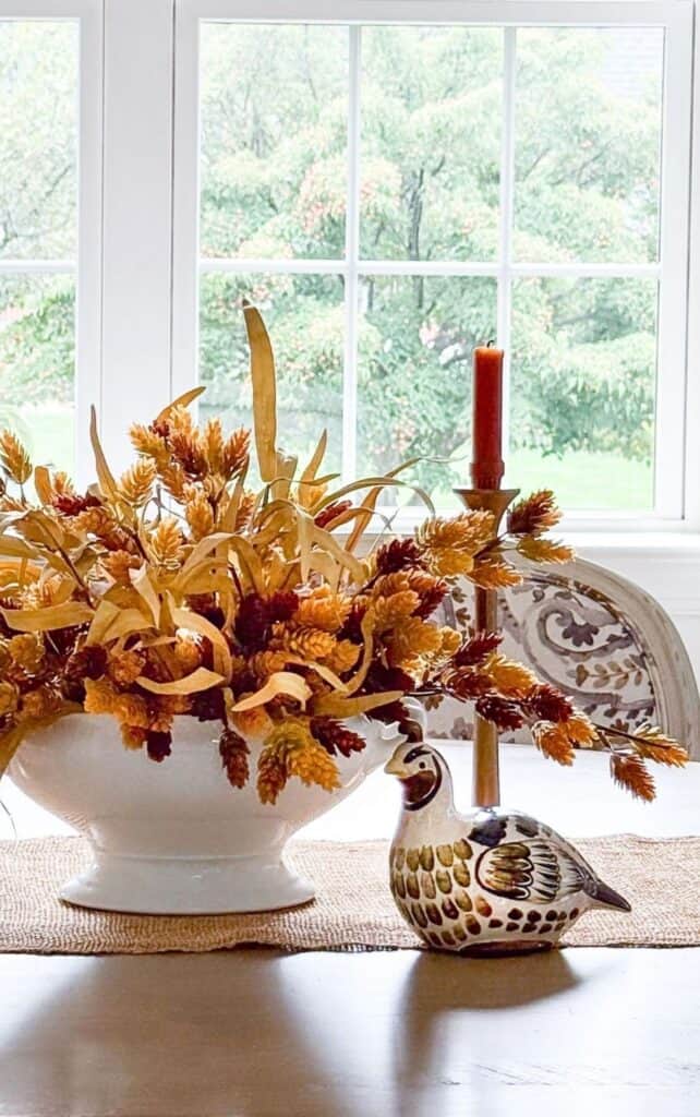 fall centerpiece with wheat and grasses in a white bowl, red taper candles, and a ceramic quail under a woven pendant light
