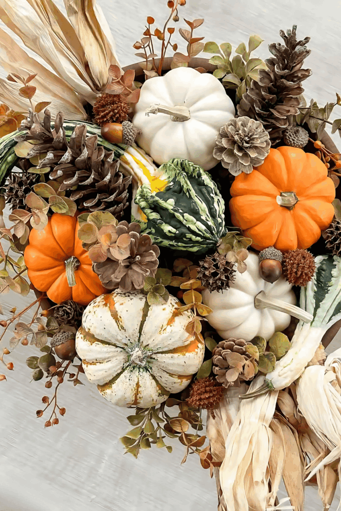 Wood dough bowl filled with pinecones, mini pumpkins, and gourds beside a candle and white pumpkins
