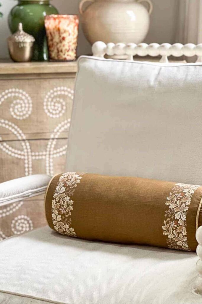 An embroidered mustard bolster pillow on a cream chair, styled with a candle and ginger jar in the background for fall.
