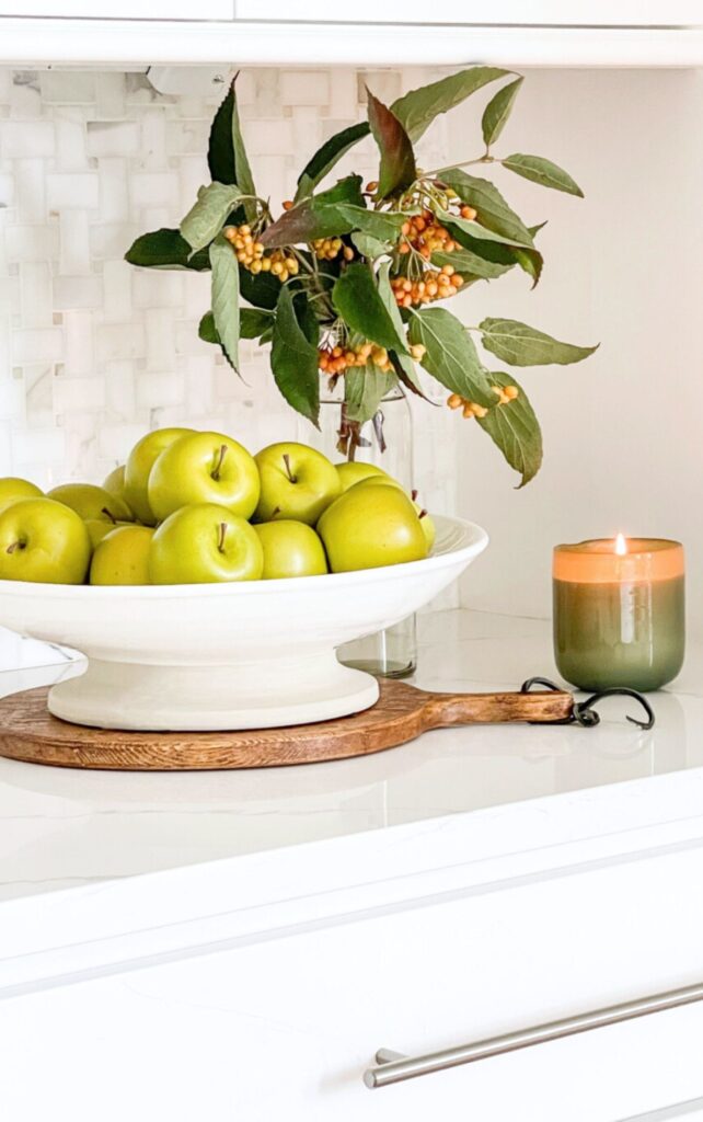 Green apples in a white pedestal bowl with a fall berry branch and lit candle on a wood board