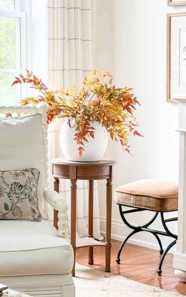 Colorful fall branches in a white vase on a wood side table beside an upholstered chair