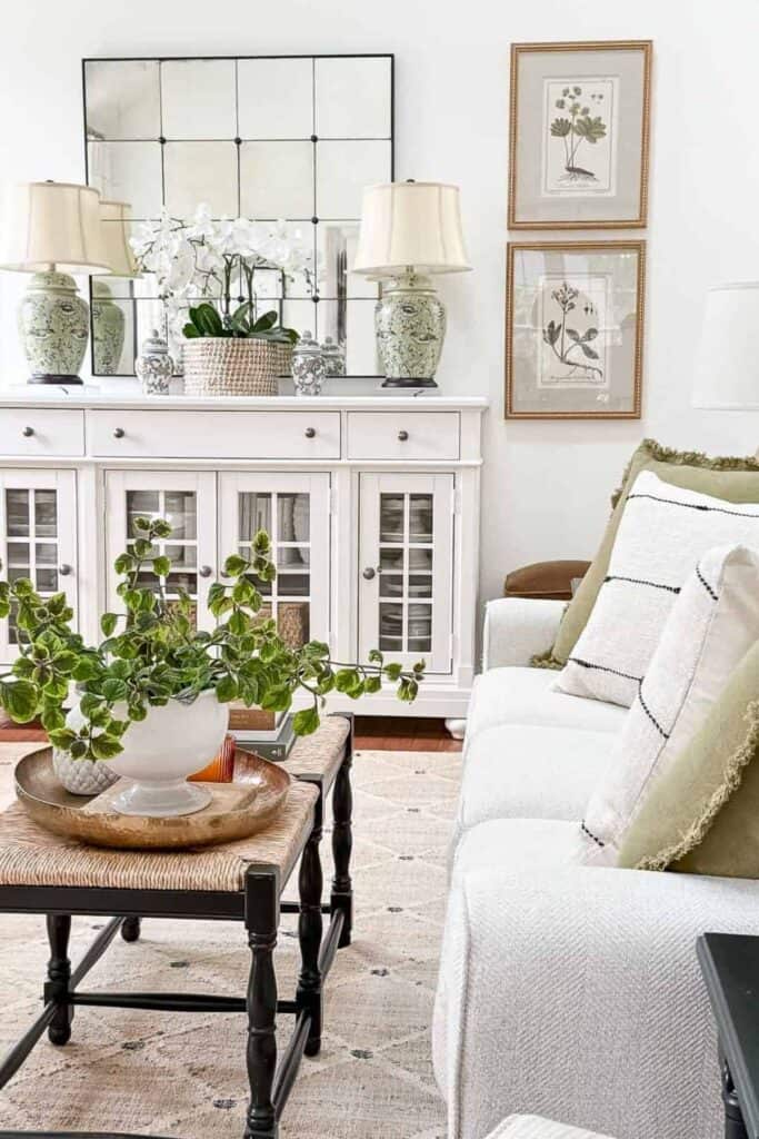 White buffet with glass front cabinets styled symmetrically with green and white ginger jar lamps, a large paneled mirror, and a white vase filled with autumn branches, beside a dark wood coffee table stacked with books.