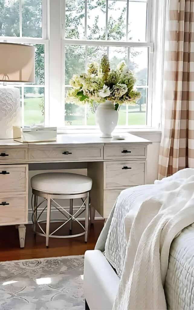 White pitcher filled with blush and green hydrangeas on a light wood vanity, styled with a small ceramic dish beside soft pink and white checked curtains.