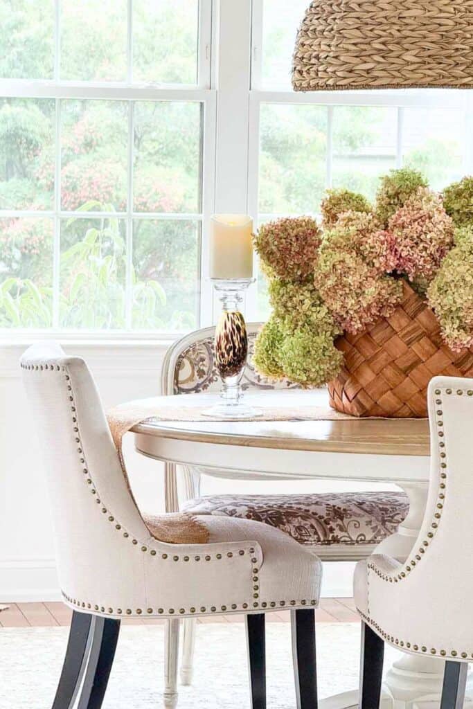 Woven basket of pink and green dried hydrangeas on a round dining table with a glass candlestick and upholstered chairs