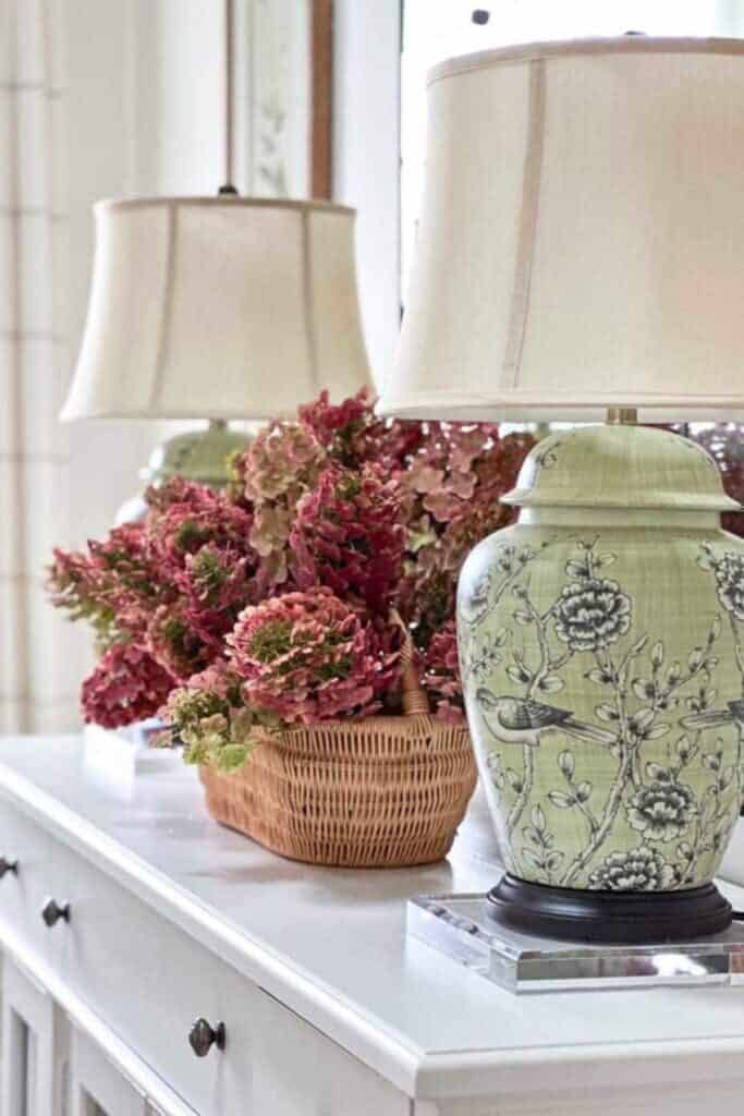 Basket of deep red hydrangeas beside green and white chinoiserie bird lamps on a white sideboard