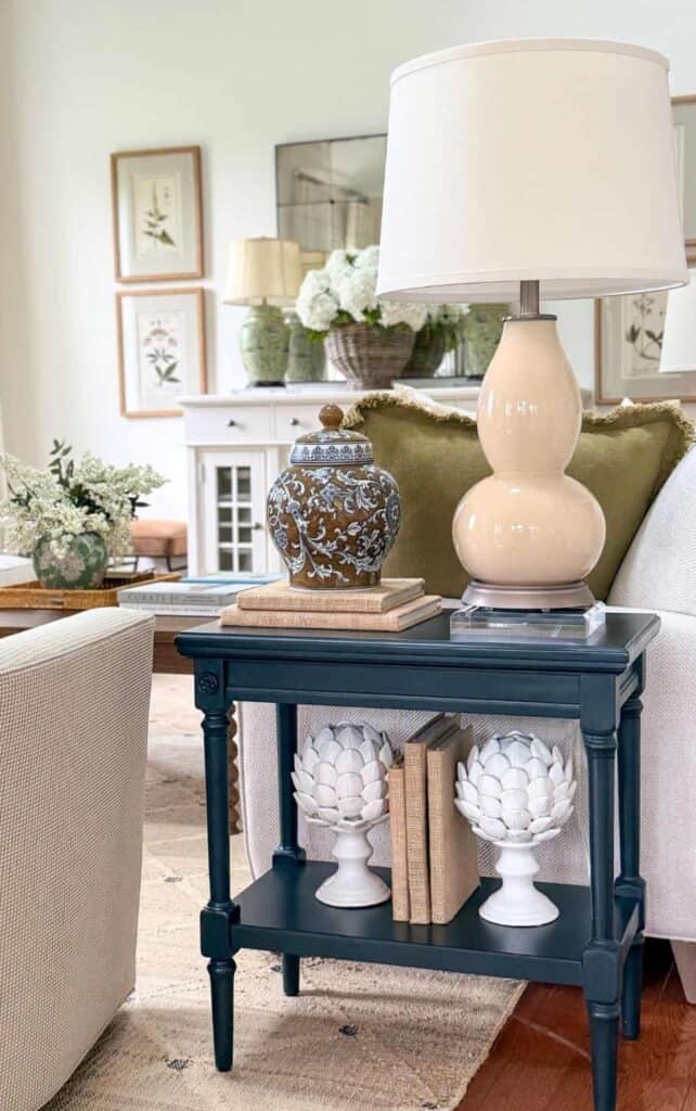 Navy blue end table styled with a double gourd lamp, blue and white ginger jar on stacked books, and ceramic artichokes on the lower shelf beside a living room sofa.