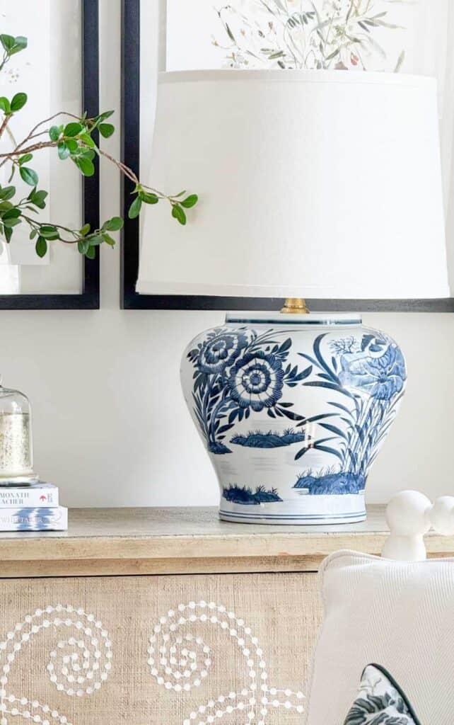 Large blue and white floral ceramic lamp with a cream shade styled on a wood dresser beside stacked books, a glass cloche, and greenery beneath framed botanical art.