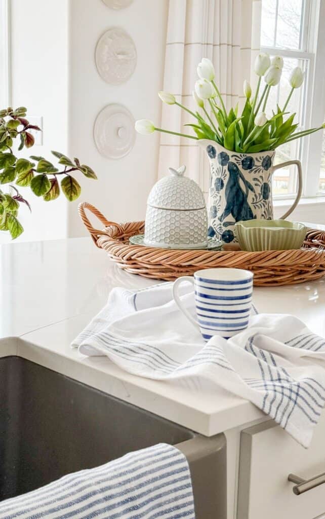 Kitchen island styled with a round woven tray holding a honeycomb butter dish, a blue and white pitcher of white tulips, a small potted pansy, and a green scalloped dish.