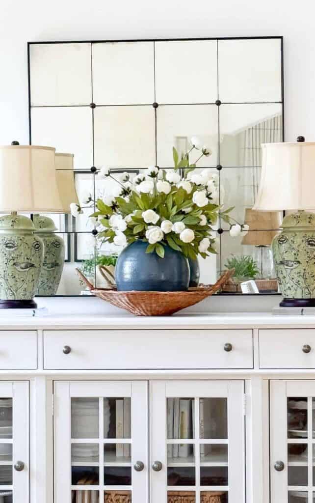 White buffet styled symmetrically with matching green and white ginger jar lamps, a large square paneled mirror, and an oversized blue vase of white blooms in a woven basket.