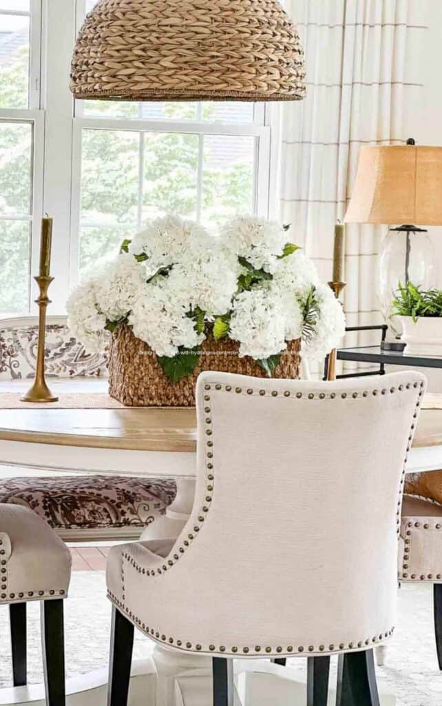 Round dining table set with an oversized white hydrangea arrangement in a woven basket, flanked by brass candlesticks, with upholstered nailhead chairs and a woven pendant light above.
