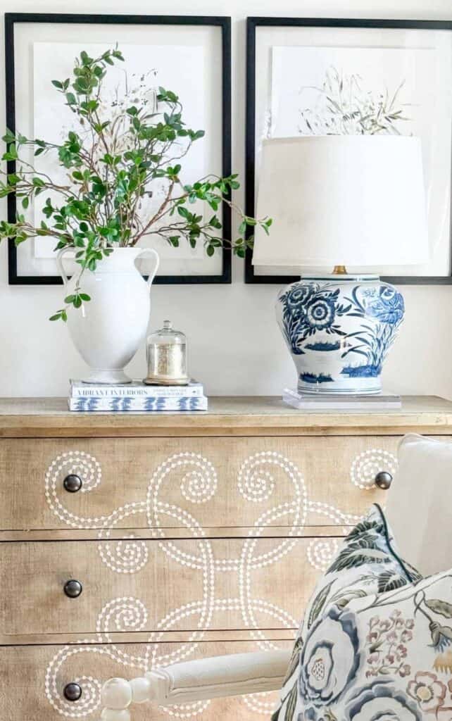Wooden dresser with stenciled swirl design styled with a blue and white floral lamp, a white pitcher of greenery, a small glass cloche, and a stack of books beneath framed botanical art.