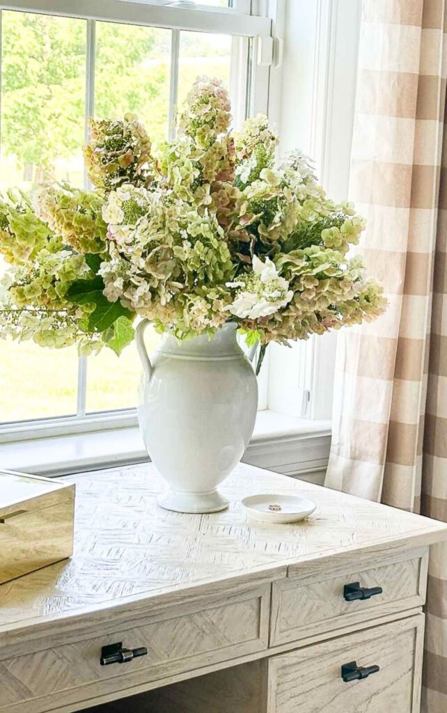 Dried hydrangeas in shades of green and pink in a white pitcher on a desk by a window