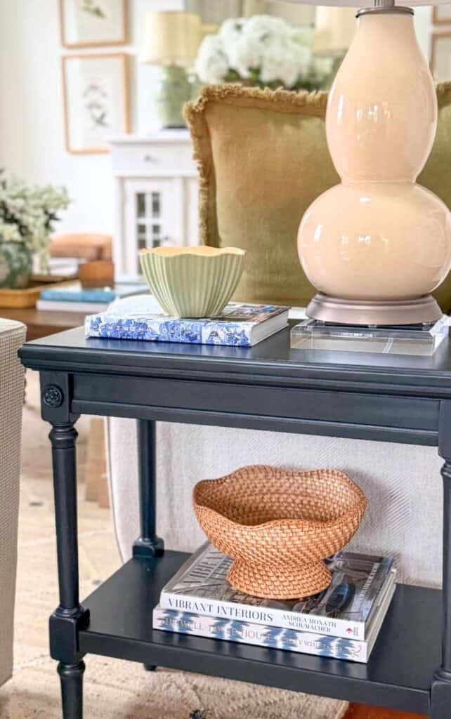 end table styled with a double gourd lamp, a scalloped green and yellow bowl on a stack of books, and a woven basket on a stack of design books on the lower shelf.