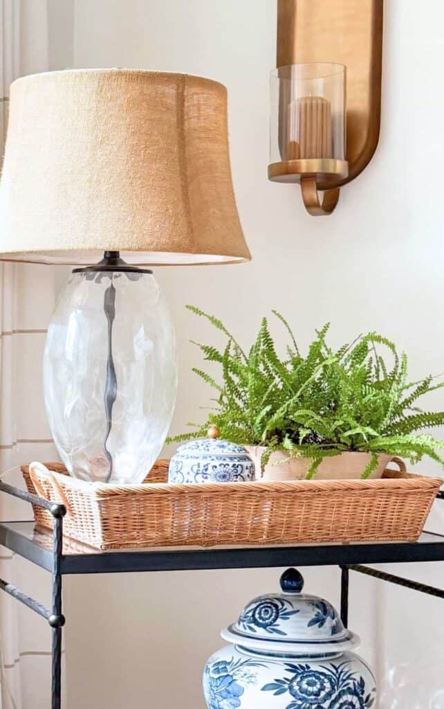 Iron console table styled with a wicker tray holding a glass lamp with a burlap shade, a blue and white ginger jar, and a fern, beneath a brass wall sconce with a candle.